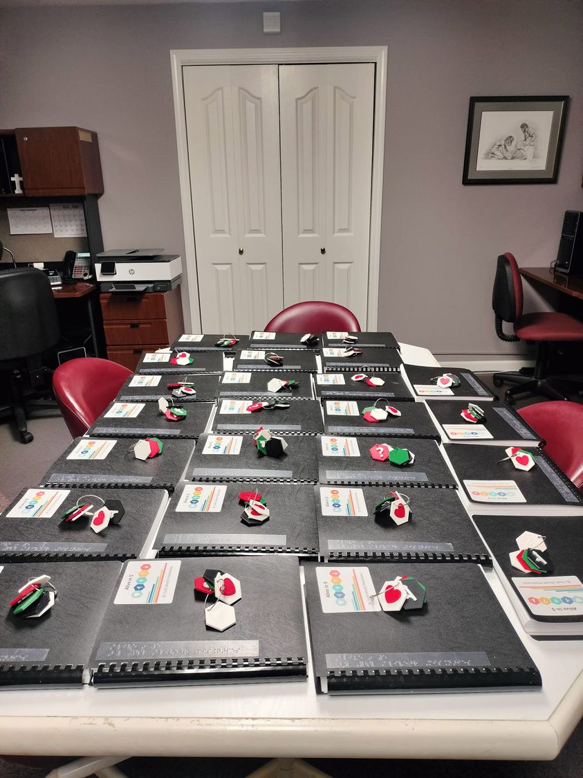 Conference table covered in black braille Alive in Five evangelistic books with 3D printed tactile keychain on top of each