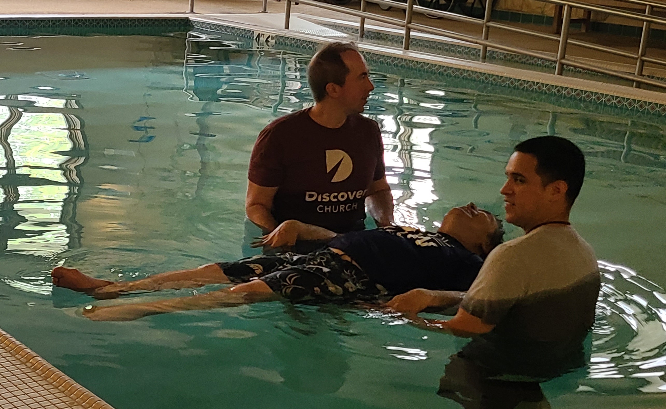 John receiving baptism in pool at local day facility pictured with two pastors