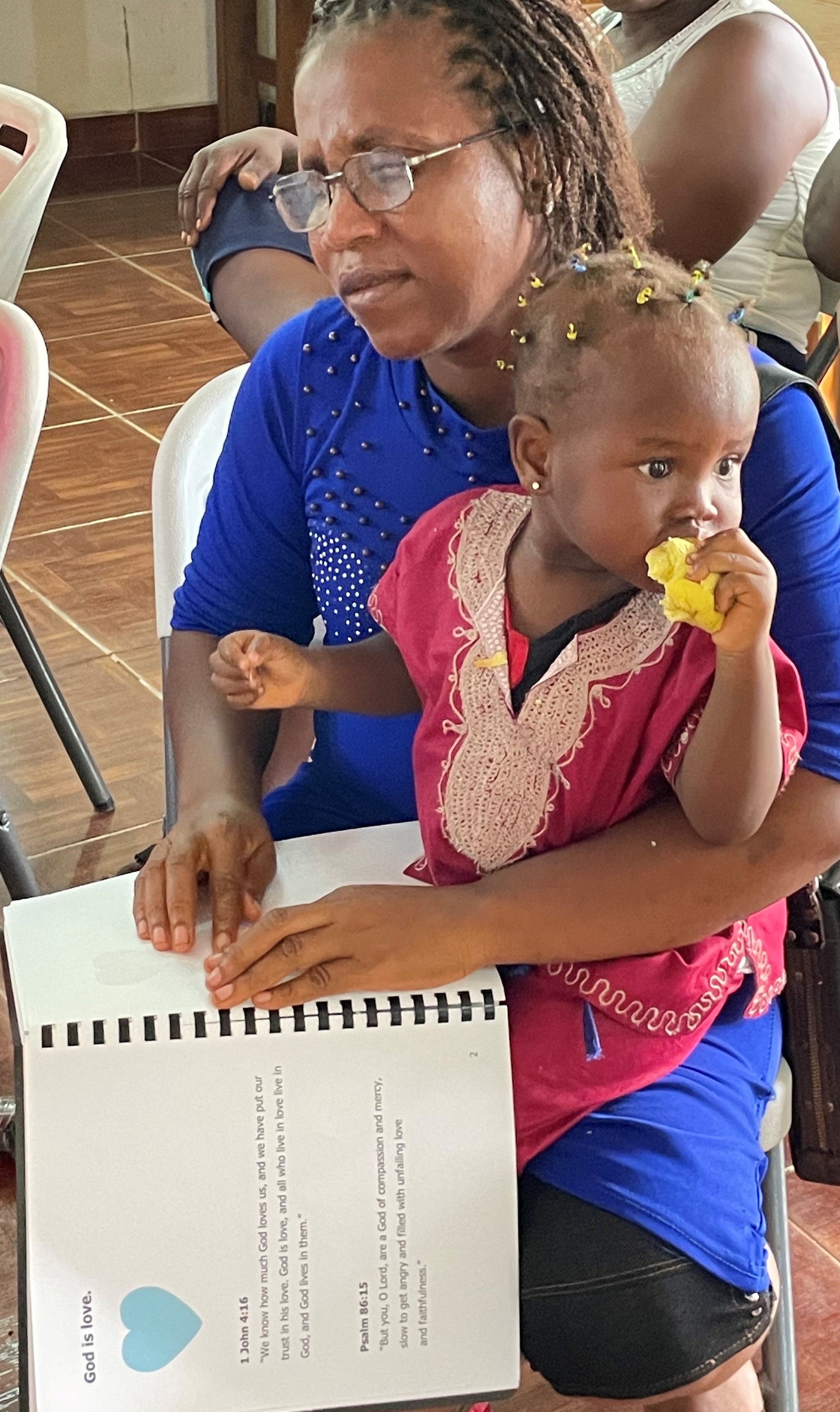 A woman holding a little girl reading the Braille Alive in Five page saying God is Love