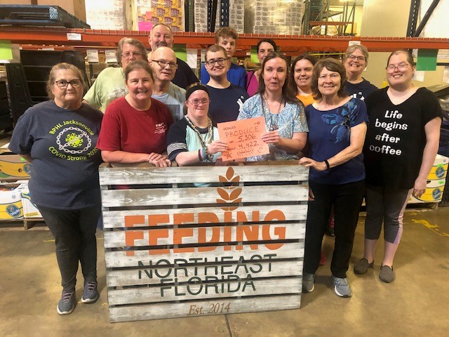 A group of people some with disabilities and some without standing smiling behind a sign reading Feeding Northeast Florida